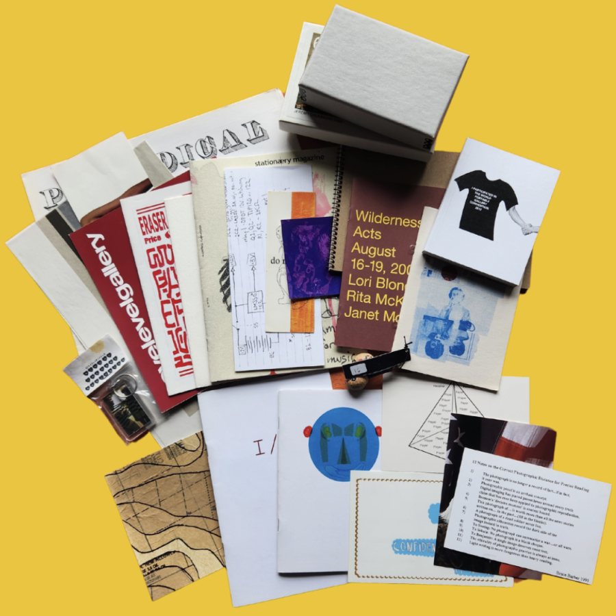 On a yellow background, contents of one of the boxes are splayed out. There is 25 items in total including, books, prints, stickers, keychains, postcards, small sculptures, original art pieces, cards, newspapers and zines. 