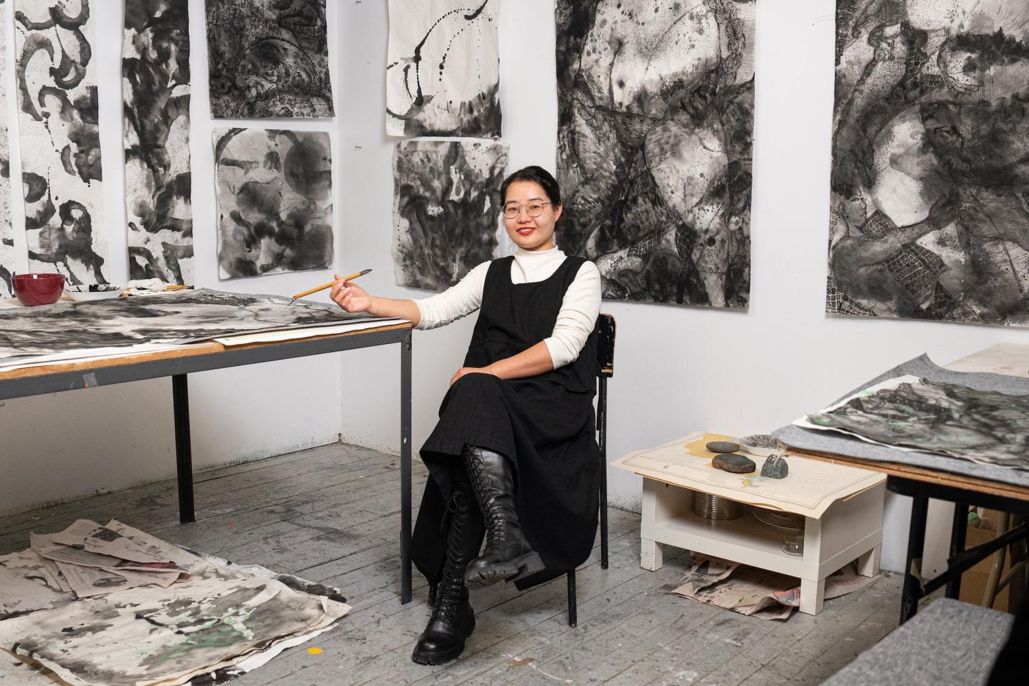ID: The artist sits in chair holding a brush, with her elbow leaning against a table. The artist is wearing a black dress, tall boots, and a white long sleeve shirt. She is wearing round glasses, and her black hair is pinned behind her head. The chair is positioned against the corner of the room, and the artists work are hung upon the walls of the room. These works are large scale paintings completed with black ink against a greyish white paper