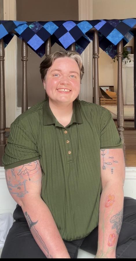 ID: a photo of Omen Mather sitting on a ledge, in front of several half finished blue quilt blocks draped over a brown bannister, smiling at the camera. Omen has light brown and grey short hair, is wearing a green short sleeve shirt, and has several tattoos on their arms.