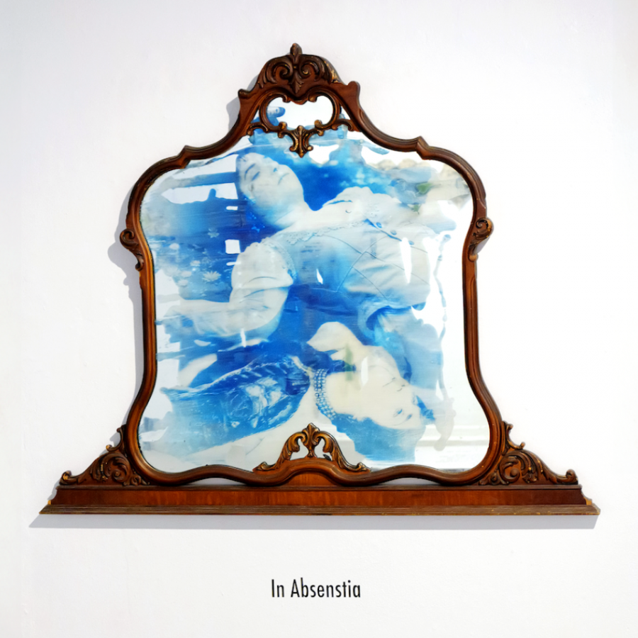 A photo of an ornamental wooden frame with a cyanotype print in it, hanging on a whitewall. The print depicts two people lying in water surrounded by floating flowers. Under the frame, large black text reads "In Absenstia".