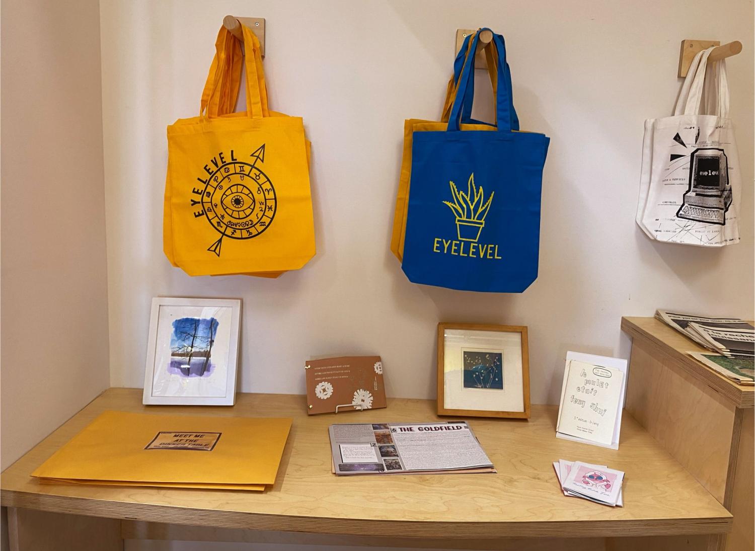 Shot of the Eyelevel Bookstore. Three types of tote bags with different designs on them, in yellow, blue, and white. Various pieces of art are organized on the tabletop.